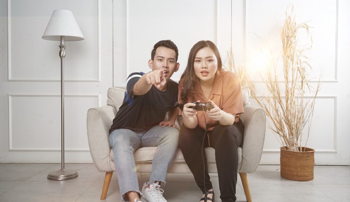 Two darker-skinned Filipinos who are sitting on a wide couch are looking at the reader. The background has a white lamp on the left and a tall plant on the right.