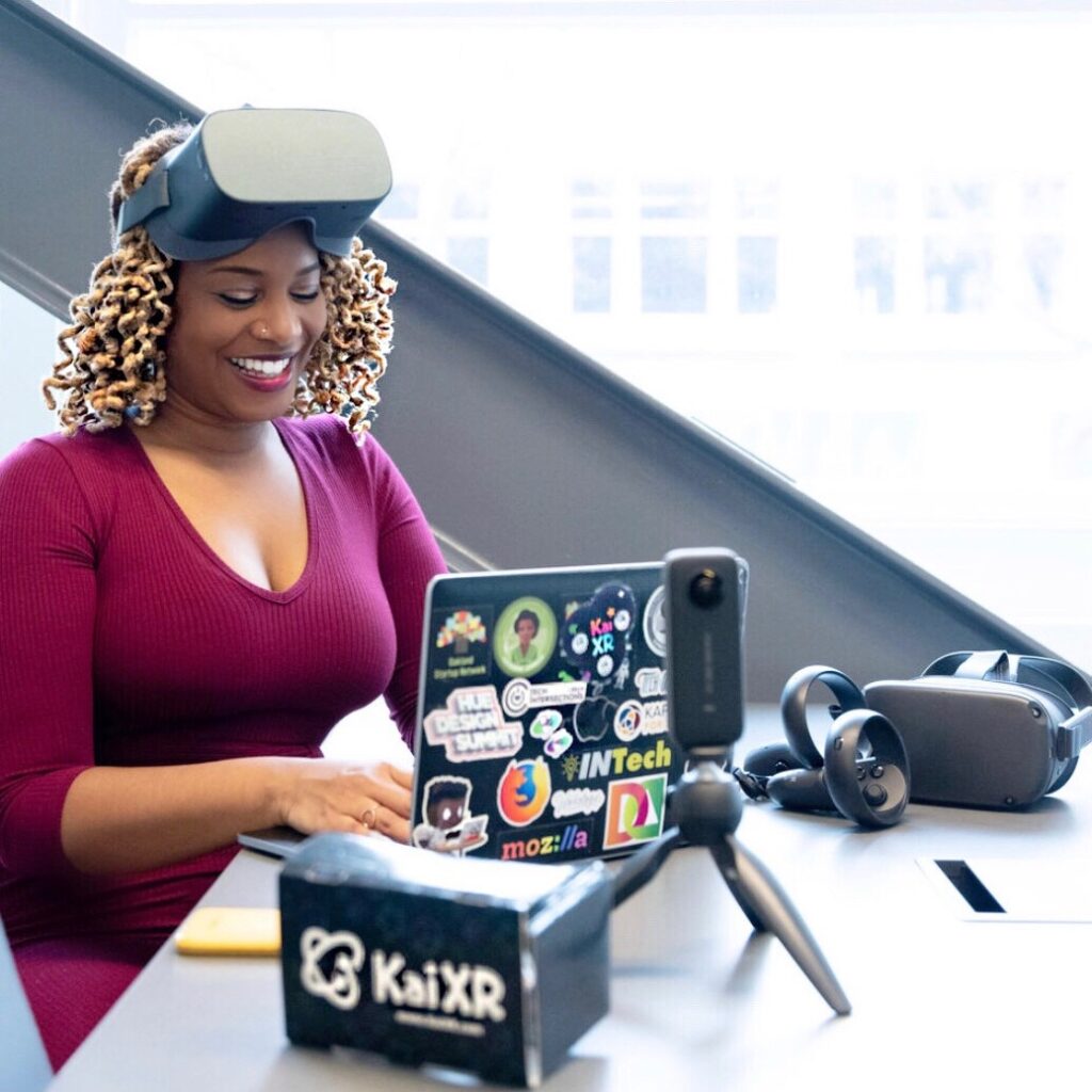 Kai Frazier - a Black woman with blonde braids using a laptop with a KaiXR headset next to her