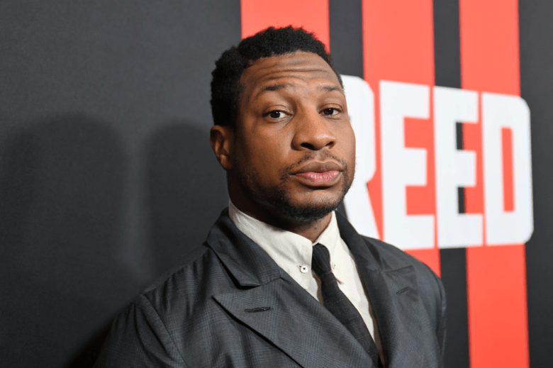 Headshot of Jonathan Majors in a suit and tie against a red and black backdrop with the words CREED spelled out in white font