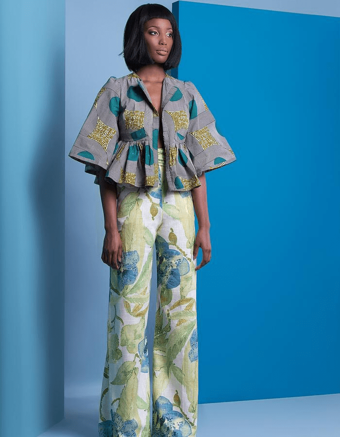 woman in shades dressed in a gray, turqouise and gold print blouse with bell sleeves and beige, green blue floral print pants standing against a turqoise background