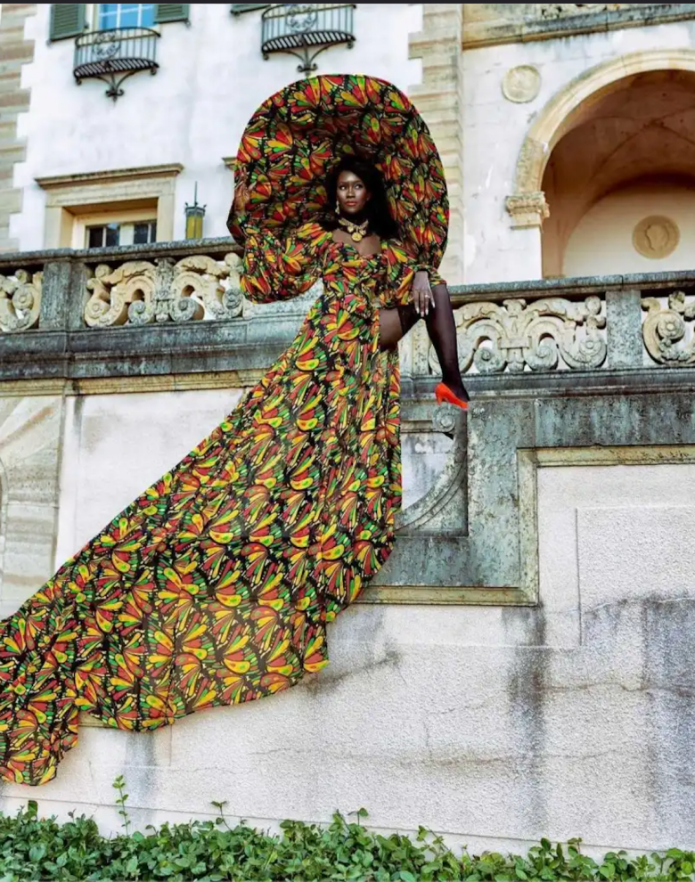 woman dressed in a red, yellow, green and black print dress with matching hat, standing outdoors on stairs